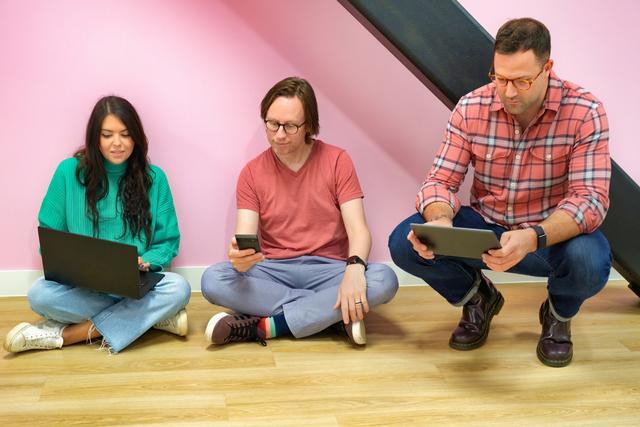 A photo of two men and one female sitting on the floor, cross-legged, looking at their laptop, phone and tablet