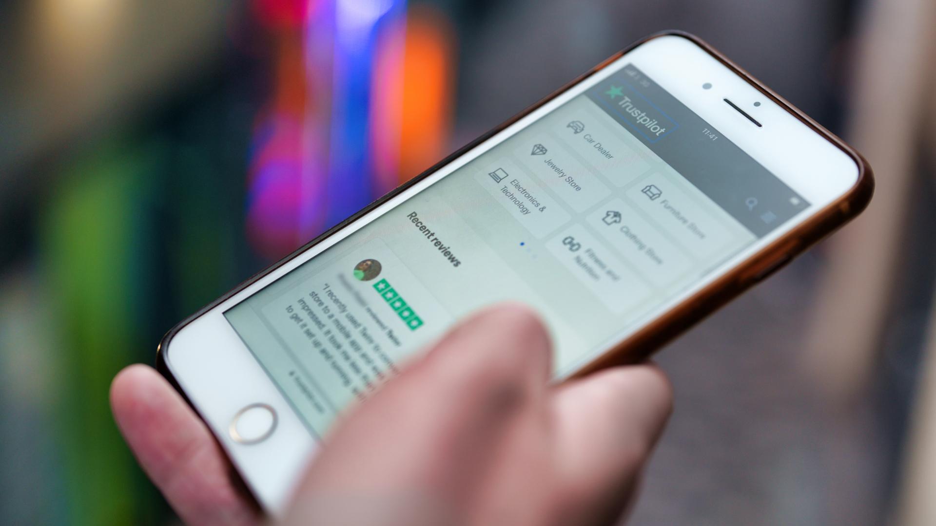 A close up photo of an iPhone in someone's hand. It is displaying the Trustpilot websites, showing recent reviews.